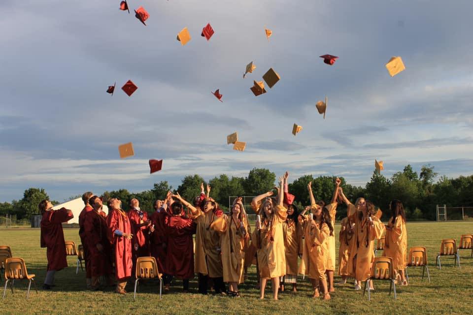 Graduating students in maroon and gold caps and gowns celebrating outdoors, tossing their caps into the air at sunset.