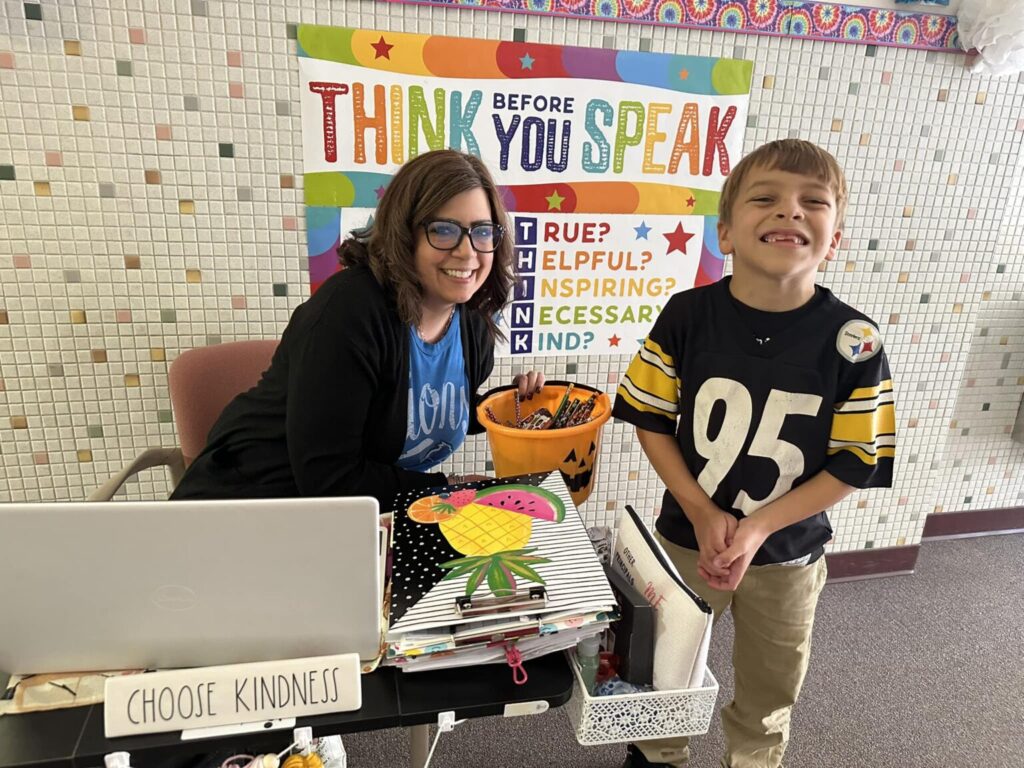 Kennedy Catholic teacher and student smile together by a desk decorated with a ‘Choose Kindness’ sign and a colorful poster reminding students to THINK before they speak.