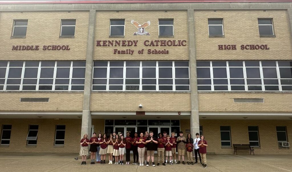 Group of middle school students standing in front of Kennedy Catholic Family of Schools building, posing together in matching maroon uniforms