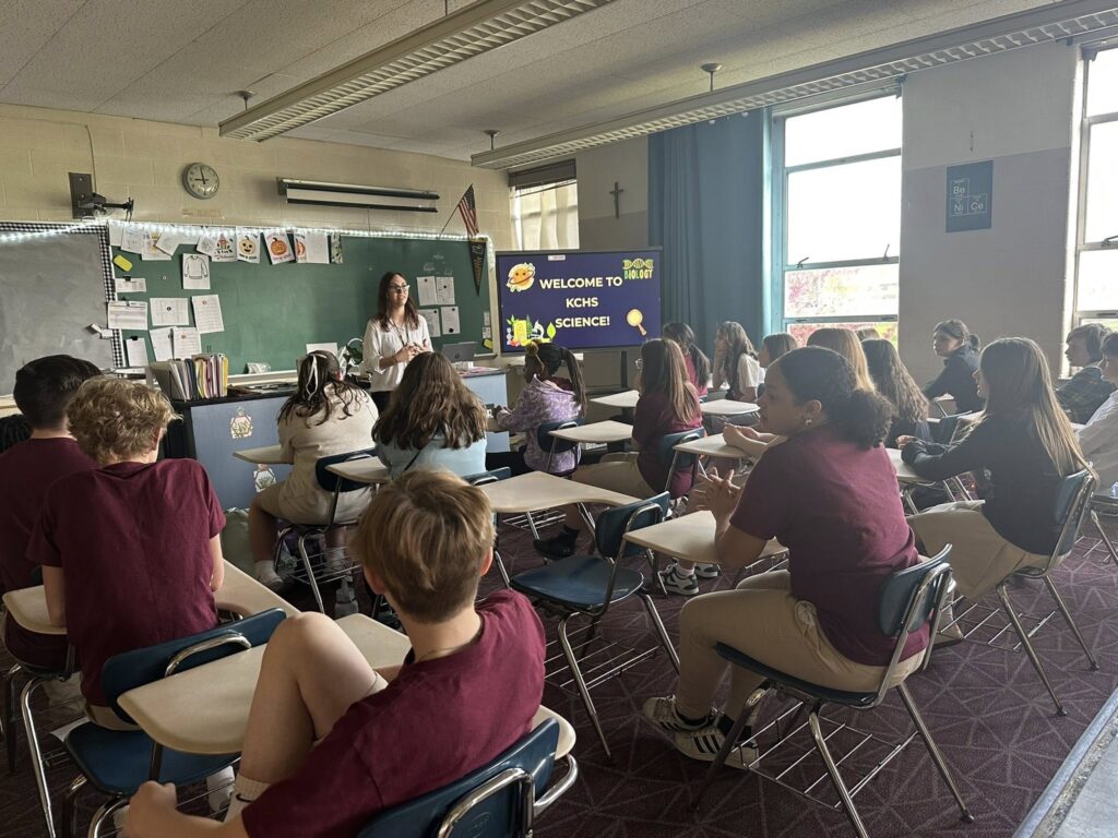 Kennedy Catholic students in maroon uniforms sit at desks while a teacher leads a science lesson with a welcome presentation displayed on the board.