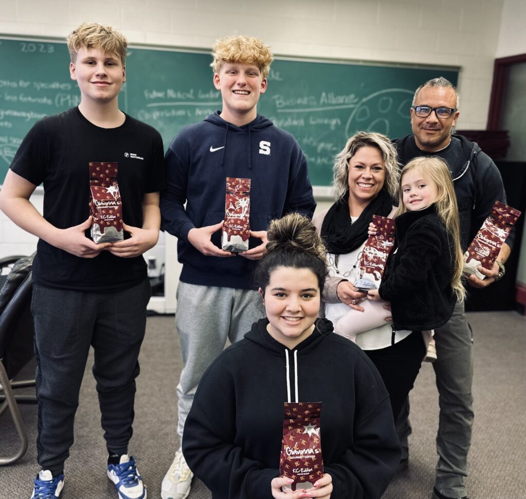 Students and family members smiling while holding Kennedy Catholic fundraiser items in a classroom setting.
