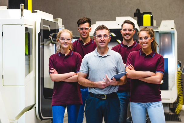 Students in safety glasses and matching uniforms stand with an instructor in front of industrial equipment in a workshop or lab setting.