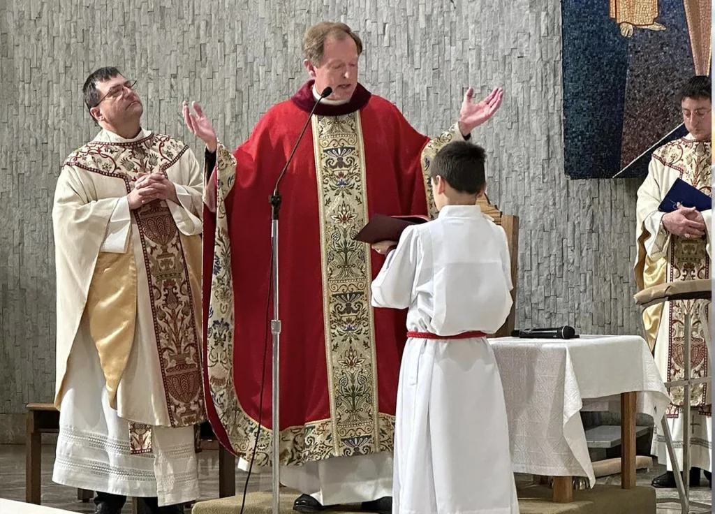 Priests and altar server during a Catholic Mass, with the celebrant in red vestments raising his hands in prayer at the altar.
